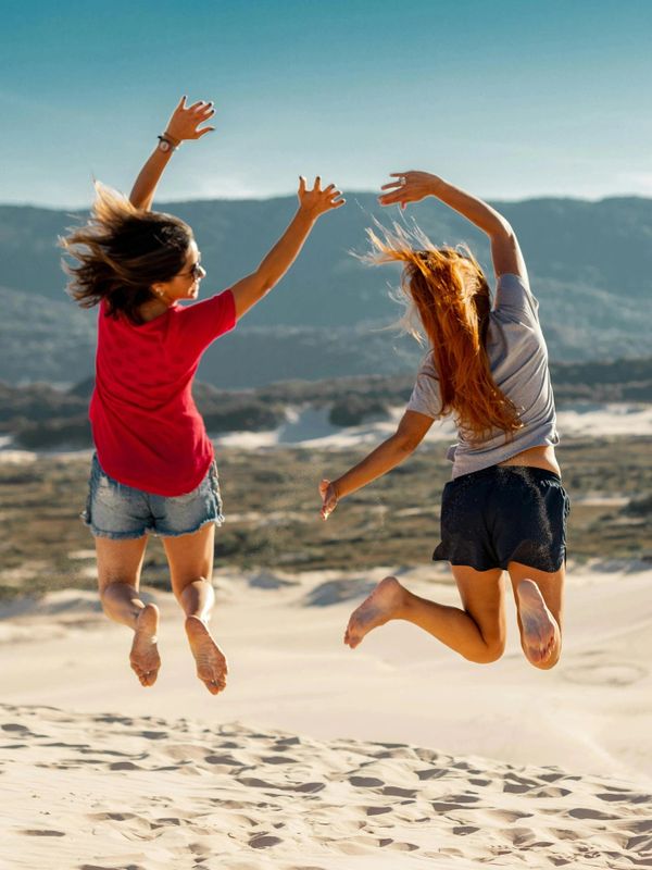 Two women jump joyfully on sand dunes with mountains in the background.