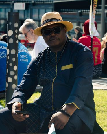 Man in stylish outfit and hat sitting outdoors with phone.
