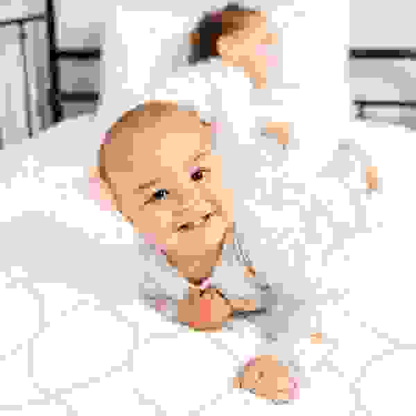 Baby lying on bed smiling at the camera with sibling in the background, illinois sleep consultant, p