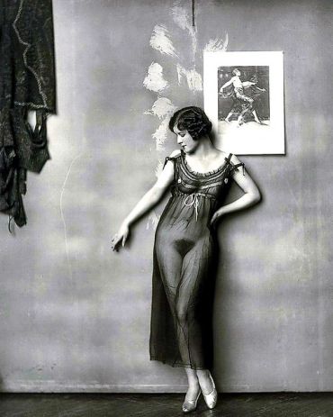 1920s woman in sheer dress posing against a wall with artwork.