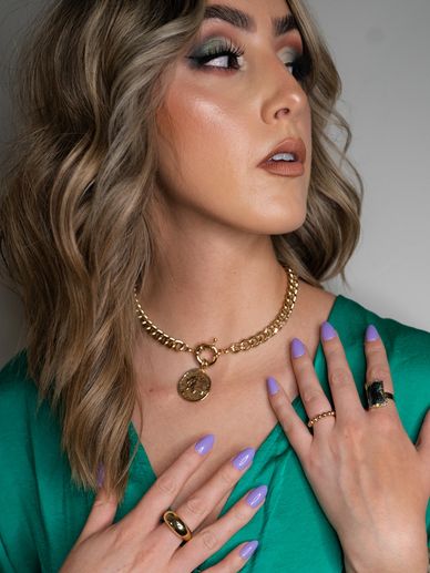 Woman with wavy hair wearing gold jewelry and lavender nails in a green top.