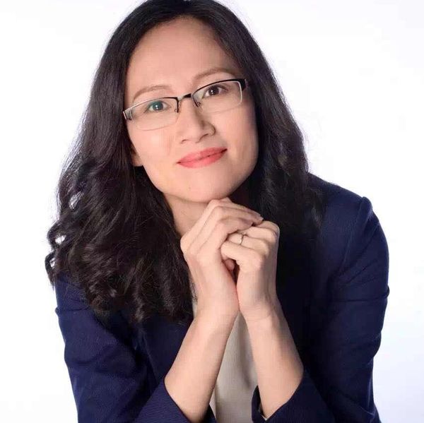 Woman with glasses smiling gently, hands clasped, wearing a navy blazer.