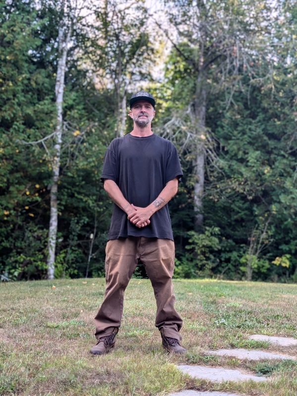 Man standing outdoors in casual attire with trees in the background.