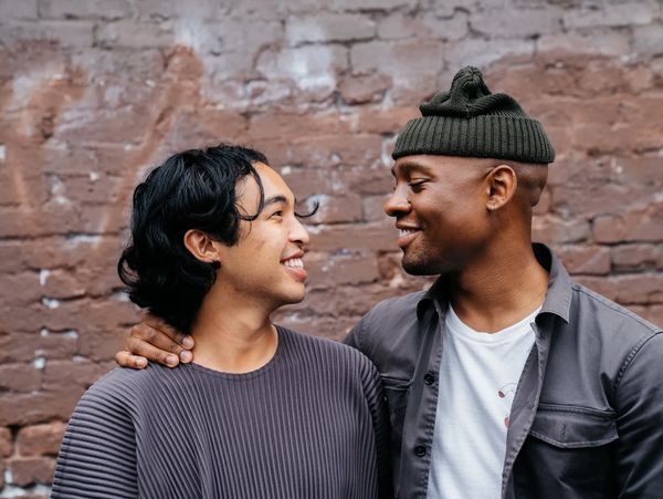 Two people smile at each other against a brick wall. One has their arm around the other's neck