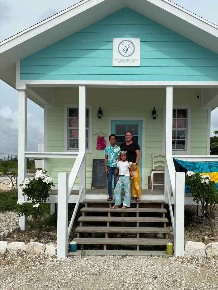 Three people standing on the porch of a colorful small shop with a turquoise and mint green facade.