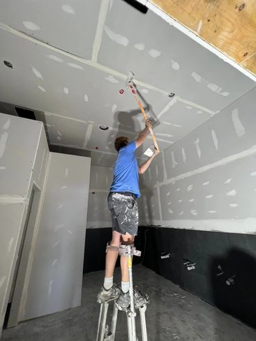 A worker on stilts applying drywall mud to a ceiling.