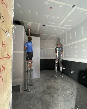 Two workers on stilts finishing drywall and ceiling joint work in a room under construction.