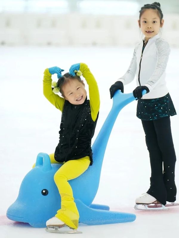 Two girls ice skating, one sitting on a blue dolphin skate aid.