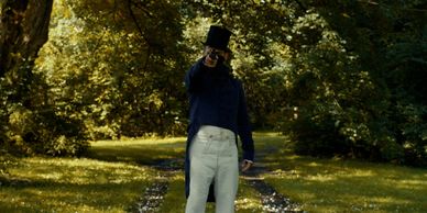 Man in vintage attire pointing a gun in a sunlit forest path.