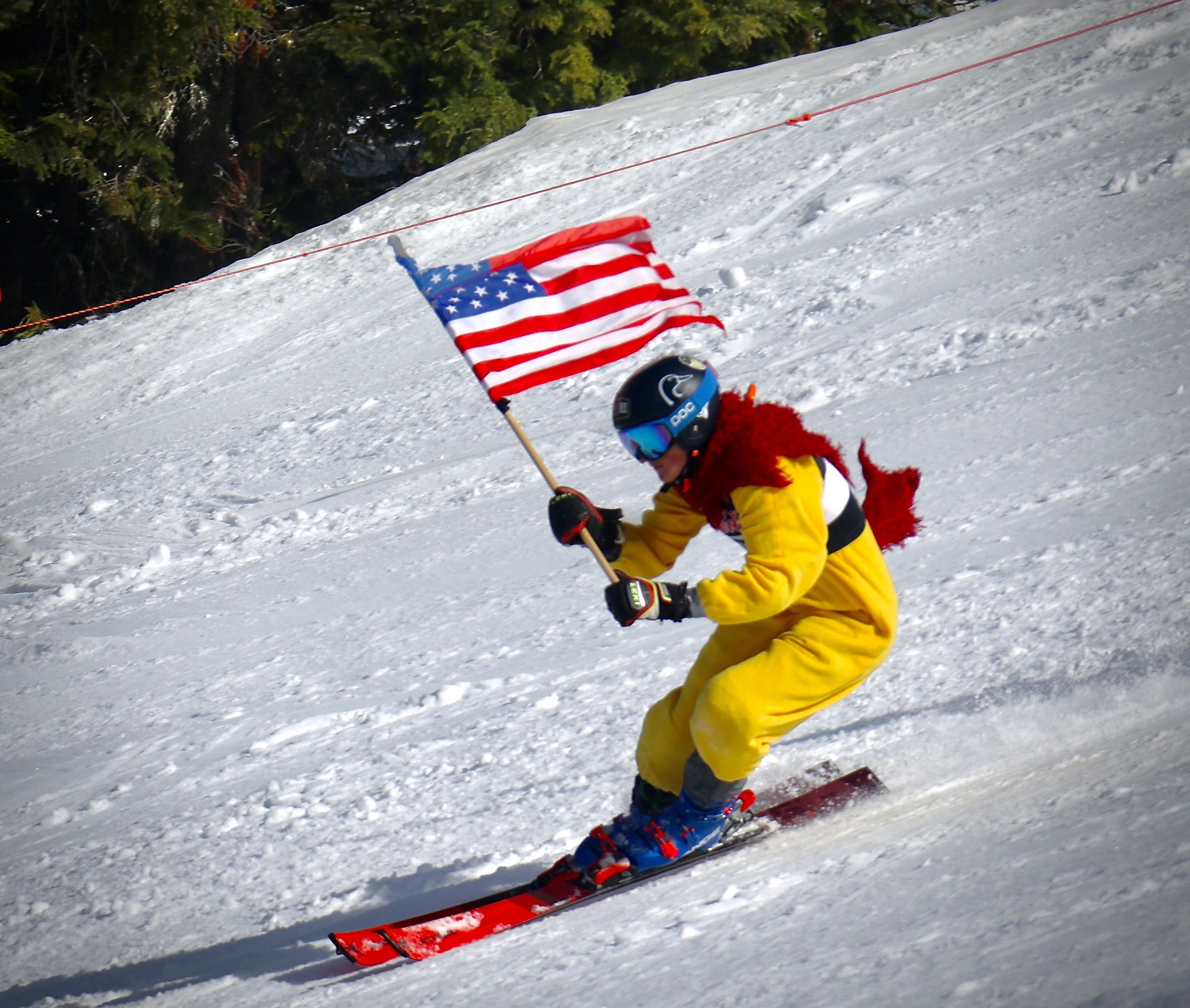 Skier in yellow chicken costume holding an American flag on a snowy slope.