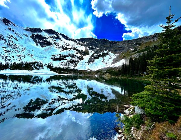 Snowy mountain reflected in a clear alpine lake under a partly cloudy sky.