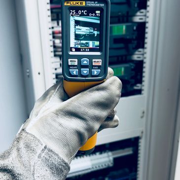 Hand holding a Fluke visual IR thermometer measuring temperature in an electrical panel.