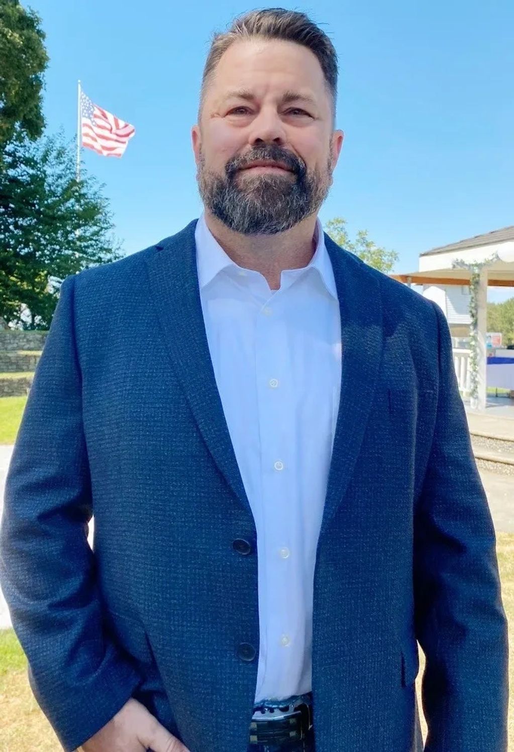 Man in a blue blazer stands outdoors with an American flag behind him.