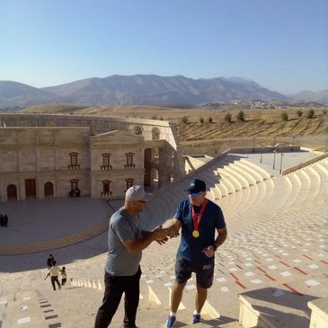 Two men shaking hands in an ancient amphitheater with mountains in the background.