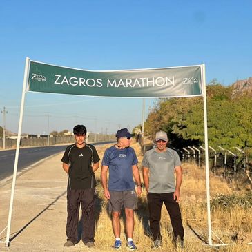 Three men standing under a Zagros Marathon banner on a sunny day.