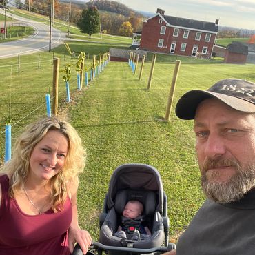 A couple with a baby stroller on a grassy hill near a red farmhouse.
