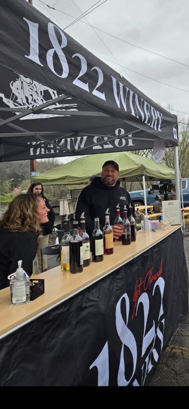 Smiling people at an 1822 Winery tasting booth with wine bottles displayed.