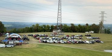 Cars parked on a grassy field near tents and power lines during early evening.