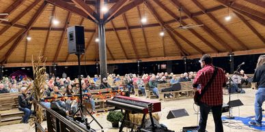 Musicians performing on stage in front of a seated audience inside a wooden pavilion.