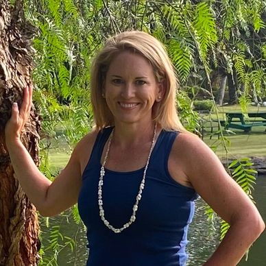 Smiling woman in a blue tank top and floral skirt stands by a tree near a pond.