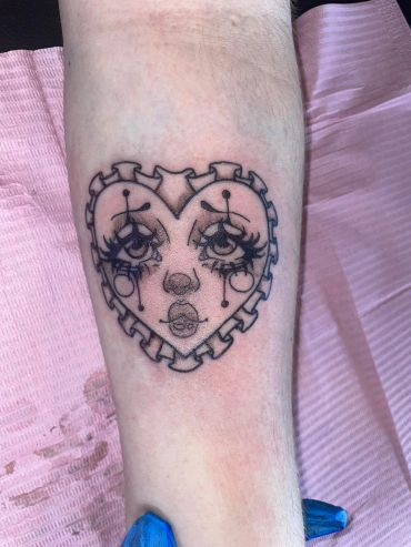 Heart-shaped sad clown face tattoo on forearm.