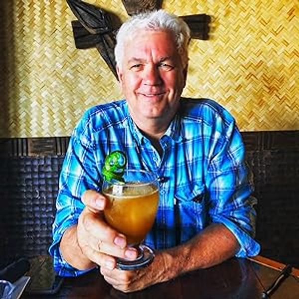 A smiling man in a blue plaid shirt holding a cocktail with a green garnish.
