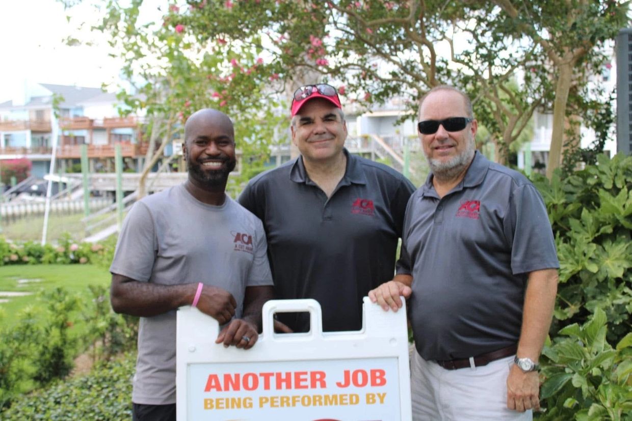 Three men smiling behind a sign outdoors.