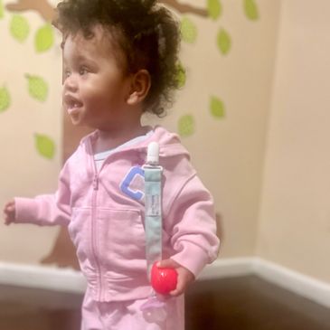 Toddler in pink outfit holding a red ball, looking to the side with a smile.