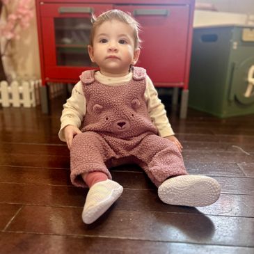 A baby in a cozy bear-themed outfit sitting on a wooden floor.