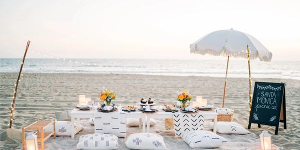 Elegant beach picnic setup with cushions, flowers, lights, and a white umbrella at sunset.