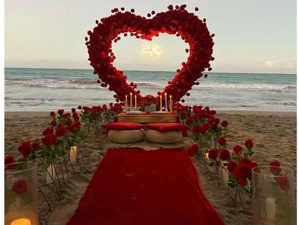 Romantic beach setup with red roses and candles under a heart-shaped rose arch.