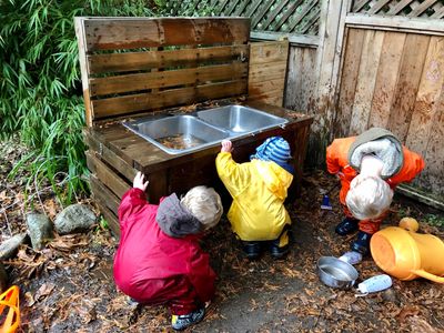 children crouch outdoors observing water flow out the underside of an outdoor sink