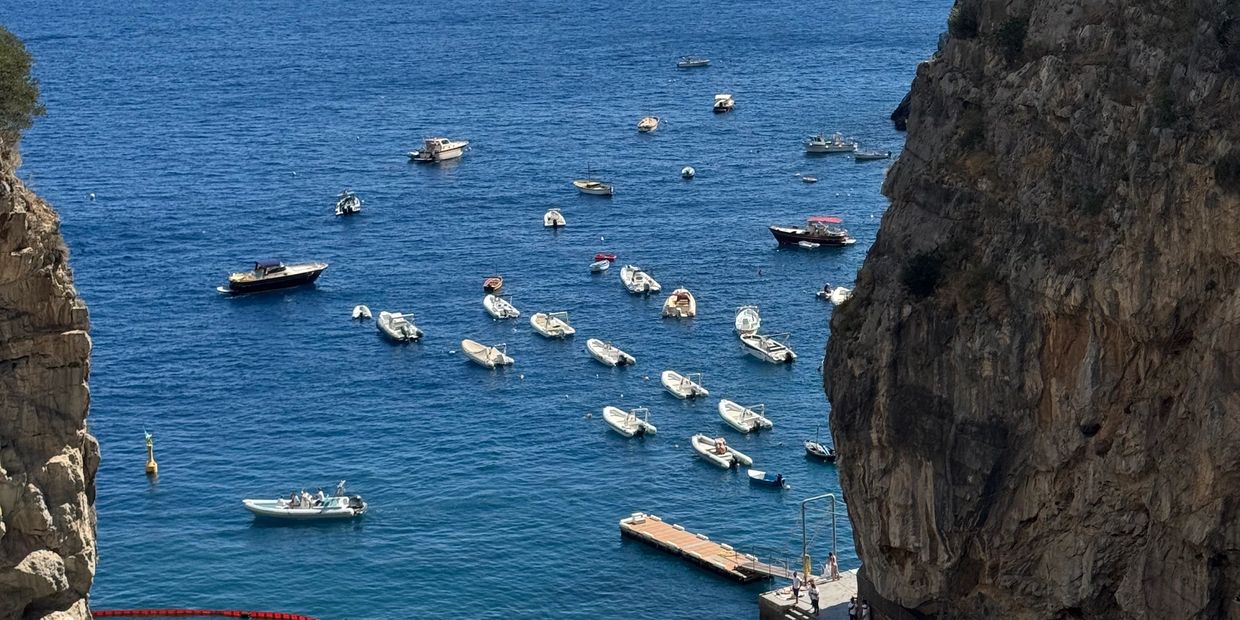 Numerous small boats anchored in calm blue waters between rocky cliffs.