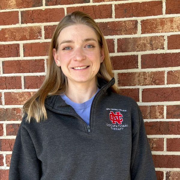 Young woman smiling in front of a brick wall wearing an occupational therapy sweatshirt.