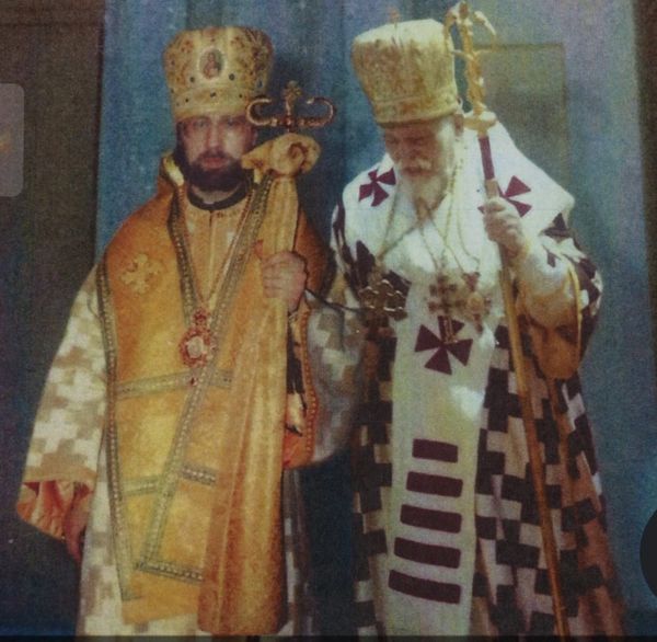 Two Orthodox clergymen in ornate religious robes and headgear hold staffs during a ceremony.