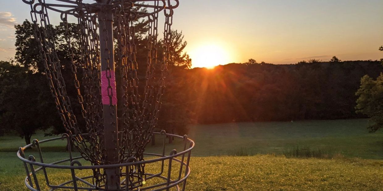 Disc golf chain basket silhouetted against a sunset over a grassy field and tree line