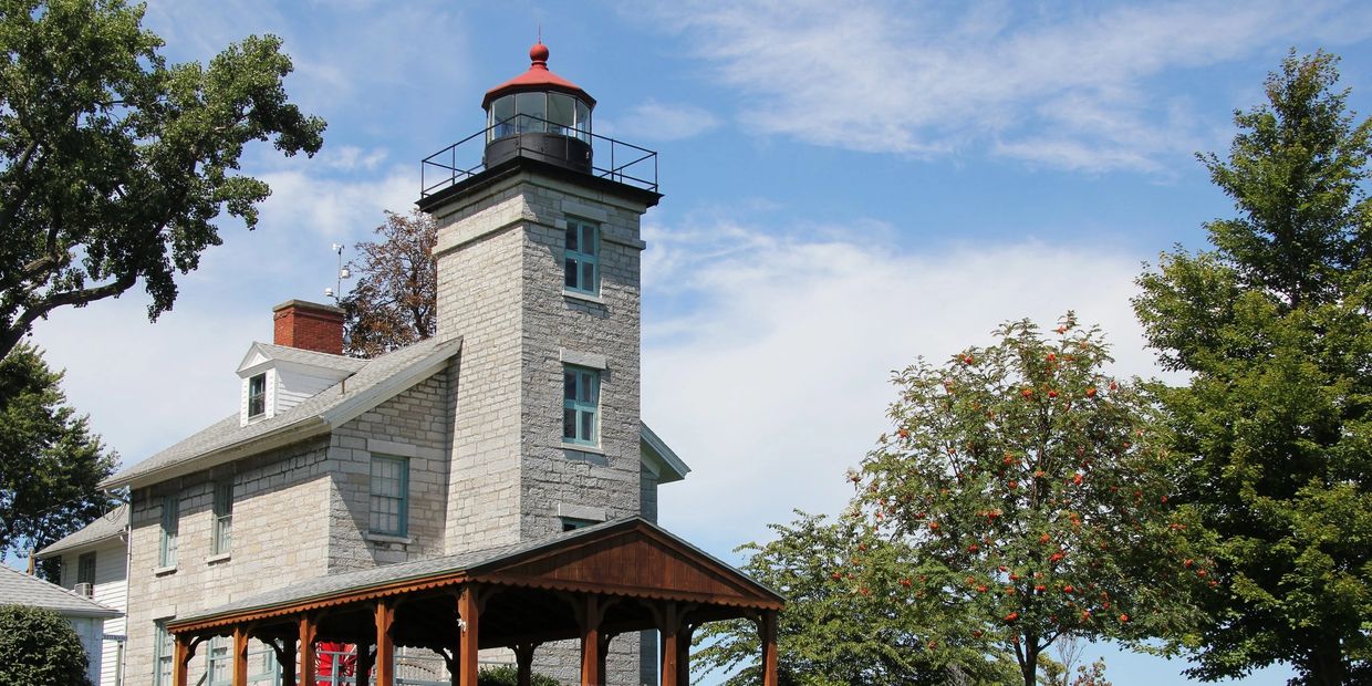 Sodus Point Lighthouse Museum, a stone lighthouse with a covered porch surrounded by green trees und