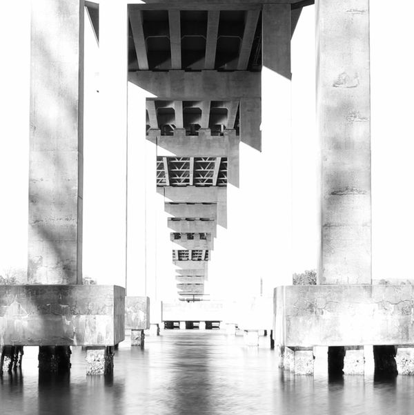Monochrome view under a concrete bridge with symmetrical pillars reflected in water.