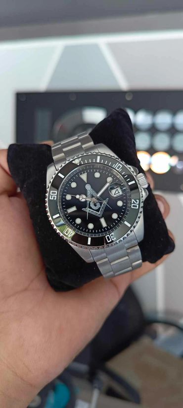 Stainless steel wristwatch with a black dial and Masonic symbol, held on a black cushion.