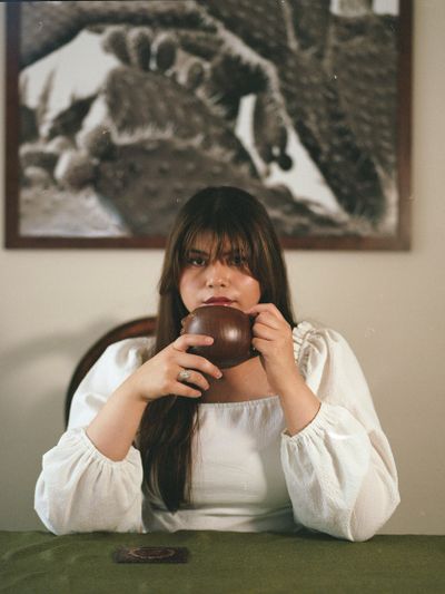 Woman in white blouse drinking from a brown cup at a table.