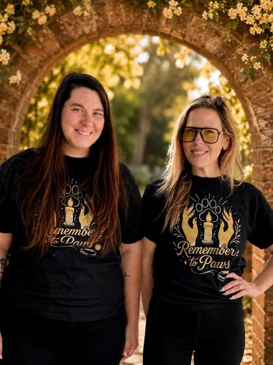 Two women wearing matching 'Remember to Paws' black t-shirts smiling under a flowered brick archway.