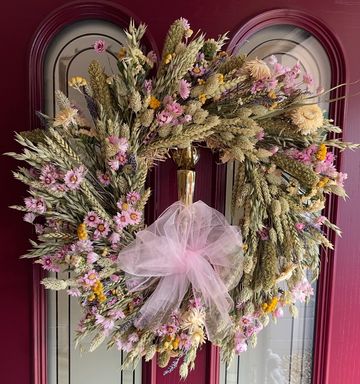 A rustic wreath made of dried flowers and wheat with a pink bow on a door.