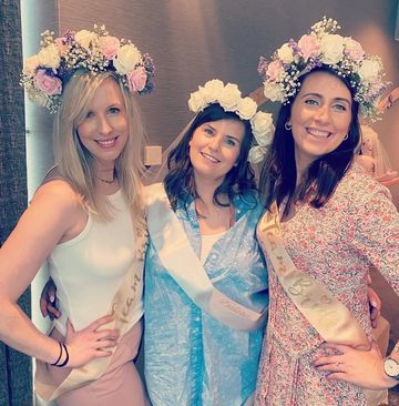 Three women wearing flower crowns and sashes, smiling for a celebration.