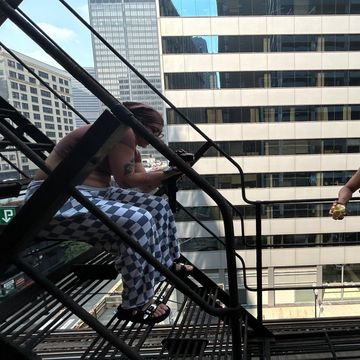 Shirtless man with checkered pants sitting on a fire escape in an urban area.