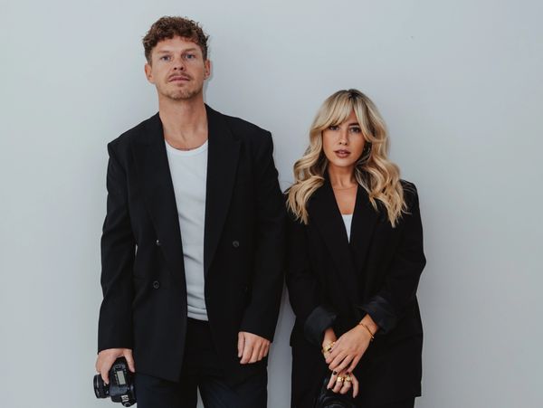 Man and woman in black blazers standing against a white wall.