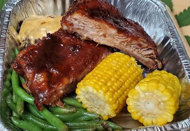 Barbecue ribs with corn on the cob, green beans, and a cheeseburger patty in a foil tray.