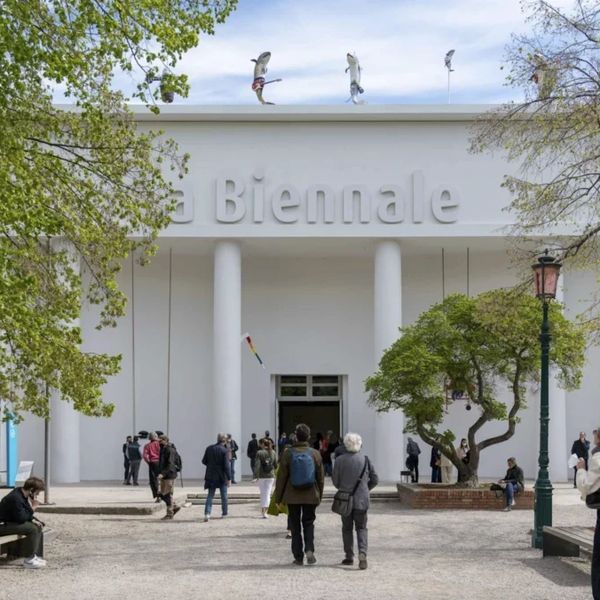 Visitors entering a white building labeled 'La Biennale' with trees and sculptures on the roof.