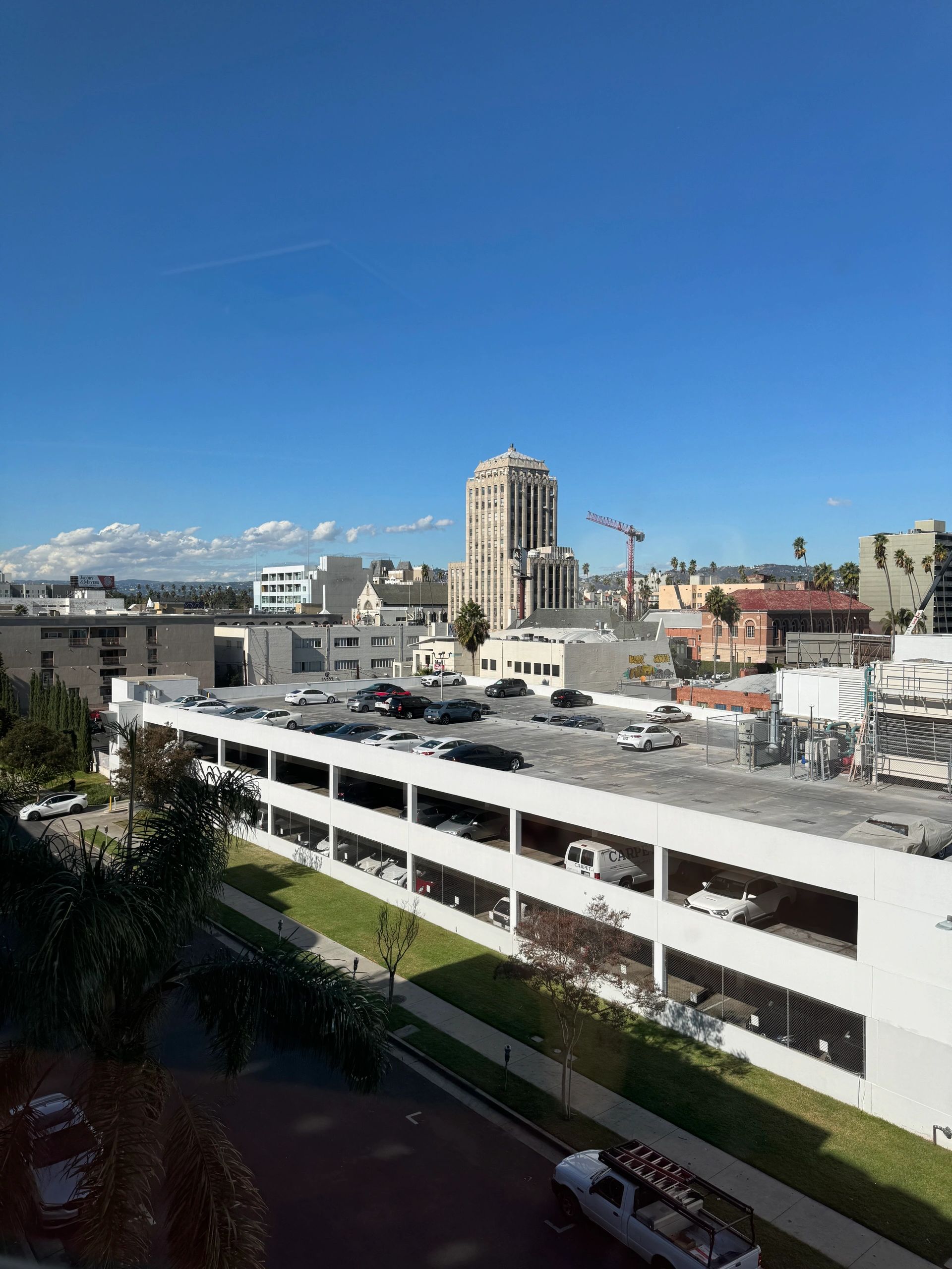 A cityscape view from a high vantage point showing a white multi-level parking garage in the foreground and various buildings in the background under a clear blue sky.