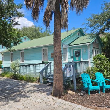 A quaint turquoise house with a green roof and two blue chairs outside.