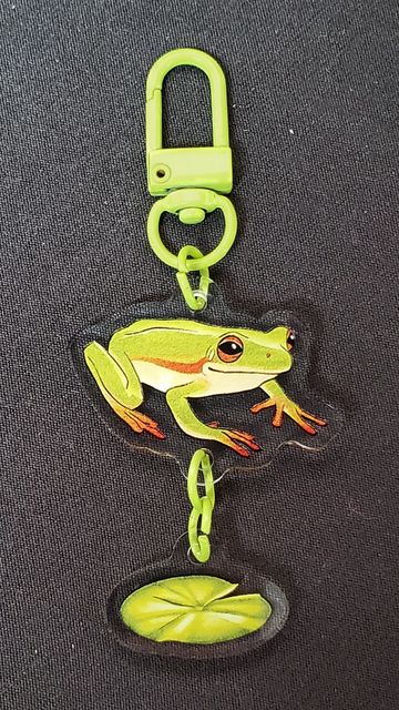 Green frog keychain with a lily pad charm on a dark fabric background.